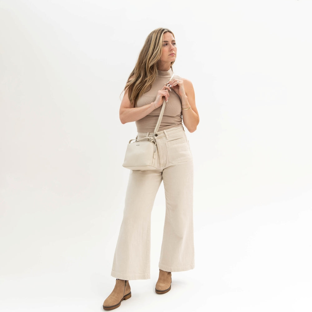 Woman wearing a beige outfit with wide-leg pants and a matching top, holding a beige handbag on a white background.