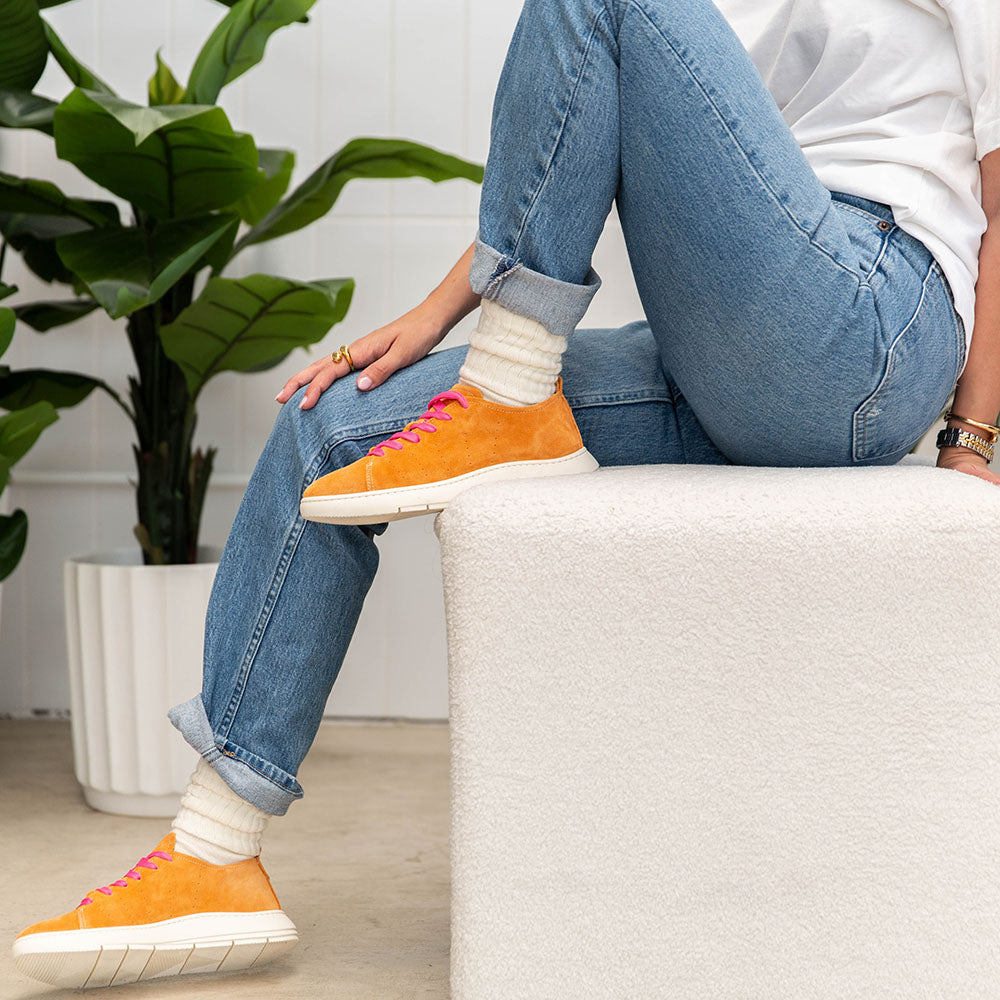 Person wearing orange sneakers and blue jeans sitting on a white surface with a plant in the background.
