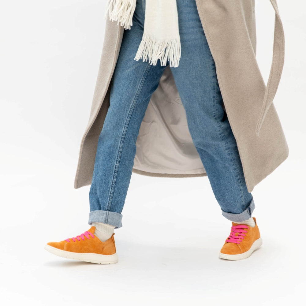 Person wearing a beige coat, blue jeans, and orange sneakers with pink laces on a white background