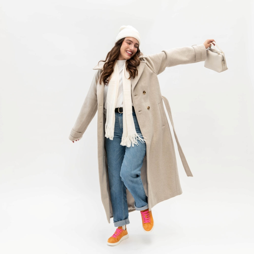 Woman wearing a beige coat, white scarf, blue jeans, and pink shoes on a white background