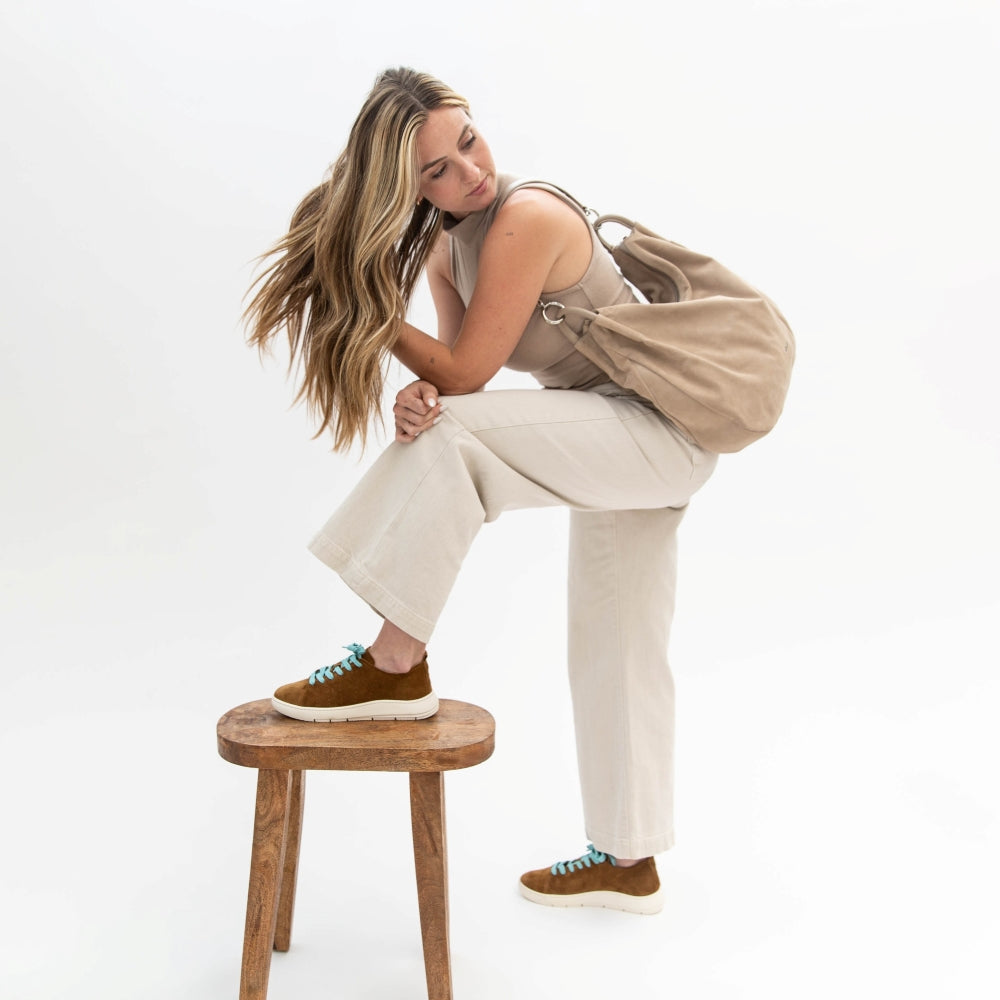 Woman sitting on a wooden stool wearing beige pants, a beige top, and brown shoes with a beige bag.