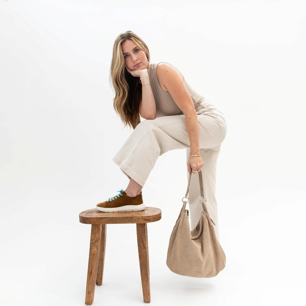 Woman sitting on a wooden stool holding a beige handbag against a white background
